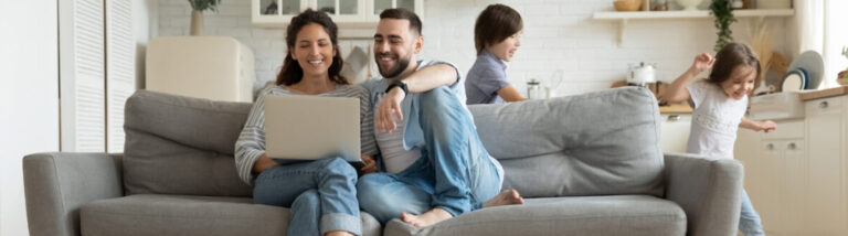 Immagine che raffigura una famiglia in casa con i genitori seduti sul divano davanti al computer, mentre i bambini giocano